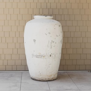 May include: A large, off-white, weathered ceramic urn with a wide body and a narrow neck. The urn has a distressed finish with visible imperfections and sits on a tiled surface. The background features a light brown, shingle-style wall.