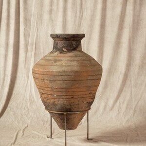 May include: An antique terracotta amphora, featuring a dark rim and a wide, bulbous body with horizontal striations. It is supported by a metal stand with three legs. The background is a neutral-toned fabric.