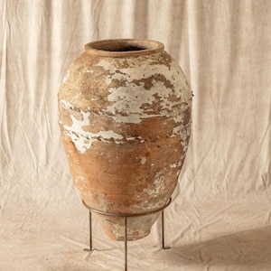 May include: A large, weathered terracotta pot with peeling white paint, set on a simple metal stand. The pot has a wide, rounded body and a slightly flared rim. The stand has three slender legs.