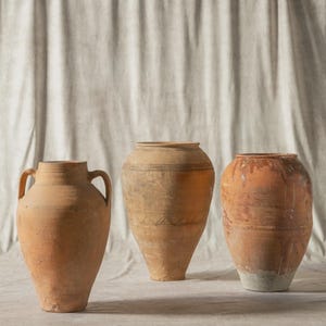 May include: Three antique terracotta vases in shades of orange-brown. The vases have different shapes and sizes, with visible texture and signs of age. One vase has handles, and another has decorative bands around the body.