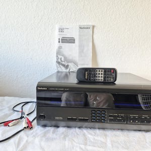 Puede incluir: Un cambiador de discos compactos Technics negro modelo SL-MC7 con un mando a distancia a juego, un manual de instrucciones impreso y un cable de audio negro con conectores RCA rojo y blanco. El panel frontal tiene una pantalla digital y botones.
