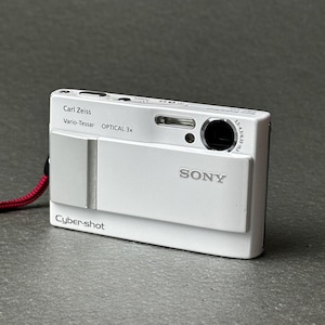 May include: A white Sony Cyber-shot digital camera featuring a sliding front cover, a Carl Zeiss Vario-Tessar lens with 3x optical zoom, a built-in flash, and a red wrist strap attached to the side. The camera body displays branding text.