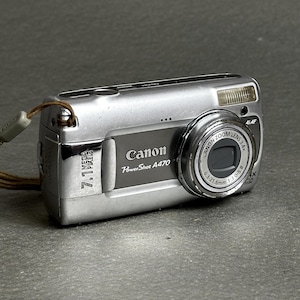 Pode incluir: Uma câmara digital prateada Canon PowerShot A470 com uma etiqueta de 7,1 megapixels, uma lente com zoom ótico de 3,4x e uma correia de pulso castanha presa na lateral. A frente apresenta um painel cinzento escuro com a marca e o modelo.
