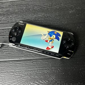 May include: A black Sony PlayStation Portable handheld gaming console featuring a wide screen displaying a graphic of the character Sonic the Hedgehog. The device includes a directional pad, action buttons, and various control buttons on the front.
