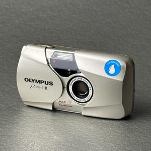 May include: Silver Olympus µ [mju:]-II camera with a 35mm lens. The camera has a built-in flash and the words "ALL WEATHER" are printed on the top. The camera is on a gray surface.