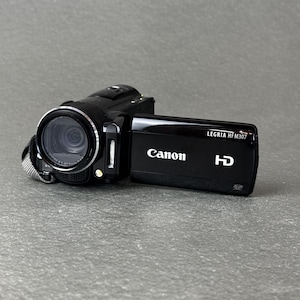 Puede incluir: Una videocámara HD Canon Legria HFM307 de color negro con la pantalla lateral abierta. El dispositivo cuenta con un objetivo circular prominente, un pequeño flash integrado y texto blanco claramente impreso en el panel lateral.