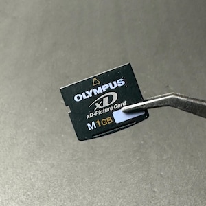 Puede incluir: Una tarjeta xD-Picture Olympus negra, sujeta con pinzas. La tarjeta tiene texto blanco que dice "OLYMPUS", "xD", "xD-Picture Card" y "M 1GB". La tarjeta es rectangular con un borde dentado.
