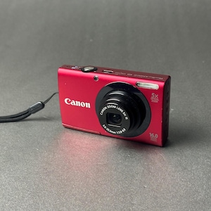 May include: A red Canon digital camera with a black lens and a wrist strap. The camera has the Canon logo and the words "5x Optical Zoom" and "16.0 Megapixels" printed on it. The lens has the words "Canon Zoom Lens 5x IS" printed on it.