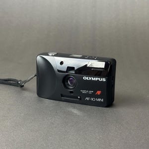 Op de afbeelding: Zwarte Olympus AF-10 Mini 35mm filmcamera met ingebouwde flitser en lens. De camera heeft een zwarte polsband bevestigd. De tekst "OLYMPUS" en "AF-10 MINI" zijn zichtbaar op de camerabehuizing.