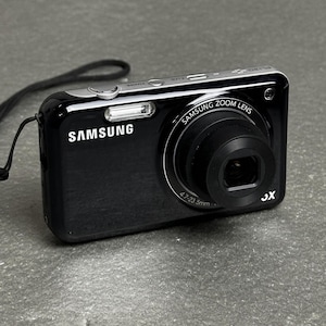 May include: A black Samsung digital camera with a silver top panel, featuring a Samsung Zoom Lens with 4.7-23.5mm focal length, a built-in flash, a power button, and a black wrist strap attached to the side of the compact rectangular body.
