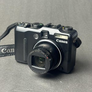 Op de afbeelding: Zwarte Canon digitale camera met zoomlens. De camera heeft een ingebouwde flitser en een riem. De lens heeft de tekst "Canon Zoom Lens 6x IS 7.4-44.4mm 1:2.8-4.8". Het Canon-logo is zichtbaar.