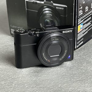 Puede incluir: Una cámara digital negra Sony Cyber-shot DSC-RX100M2 con un objetivo Carl Zeiss Vario-Sonnar T* con especificaciones 1,8-4,9/10,4-37,1. El cuerpo de la cámara incluye un dial de modo superior, un botón de disparo y un flash emergente.