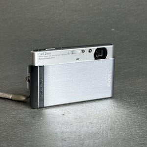 May include: A silver Sony Cyber-shot digital camera with a brushed metal finish. The front features a Carl Zeiss Vario-Tessar 3.5-4.6/6.18-24.7 lens, an optical flash, and the Sony logo. A small wrist strap is attached to the side of the device.