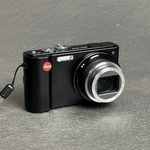 Pode incluir: Uma câmara digital preta Leica V-Lux 30 com um canhão de lente prateado com o texto DC VARIO-ELMAR 1:3.3-4.9/4.3-19.2 ASPH. O corpo da câmara inclui um logótipo vermelho da Leica, um flash integrado, um botão de disparo prateado e uma correia de pulso preta.