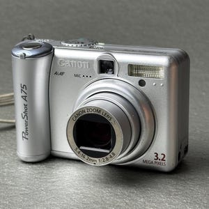 May include: Silver Canon PowerShot A75 digital camera. Features a zoom lens, built-in flash, and a 3.2-megapixel resolution. The camera has the Canon logo and PowerShot A75 model name printed on the body.