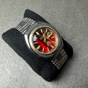 May include: A vintage wristwatch with a stainless steel strap and case. The watch face is a gradient of red and black with gold accents and the date displayed. The watch is resting on a black surface.