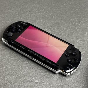 Pode incluir: Uma consola de jogos portátil Sony PSP preta com acabamento brilhante, ecrã panorâmico com fundo degradé rosa e laranja, botões direcionais, botões de ação e etiquetas de controlo incluindo Select, Start, Power e Hold.