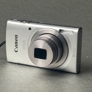 May include: A silver Canon IXUS digital camera featuring an 8x optical zoom lens. The front of the camera displays the Canon logo, the IXUS model name, and lens specifications including 5.0-40.0mm. A built-in flash is located on the top right corner.