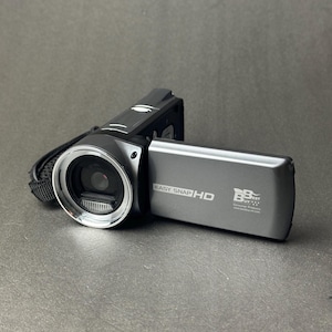 May include: A black and silver digital camcorder with the screen open. The camcorder has a lens with a silver ring around it. The screen displays the text "EASY SNAP HD" and a logo. A black wrist strap is attached to the side.