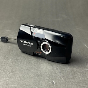 May include: Black Olympus µ [mju:]-II camera with a 35mm lens. The compact, all-weather camera has a built-in flash and a wrist strap. The camera's lens is labeled "LARGE APERTURE LENS". The camera is on a gray surface.