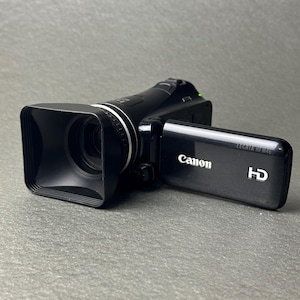 Op de afbeelding: Zwarte Canon LEGRIA HF M46 HD camcorder met een zonnekap en een uitklapbaar scherm. Het Canon-logo is zichtbaar op het scherm. De camcorder ligt op een grijze ondergrond.