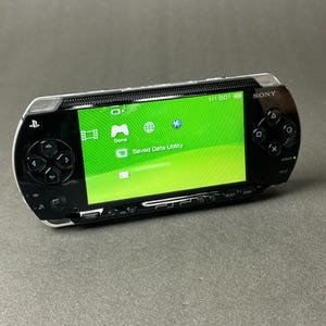 Puede incluir: Una consola de juegos portátil Sony PSP negra y plateada. La pantalla muestra un menú verde con iconos de juegos y datos guardados. La consola tiene un pad direccional, botones y un joystick.