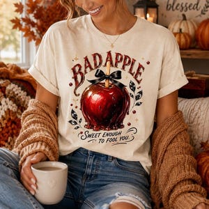 May include: A cream-colored short-sleeve t-shirt featuring a graphic of a shiny red candy apple with a black bow. The text BAD APPLE is printed in a dark red font above the apple, and the text SWEET ENOUGH TO FOOL YOU is printed below it.