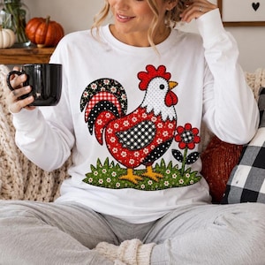 May include: A white long-sleeve shirt featuring a large graphic of a rooster composed of various red, black, and white patterned patches, including gingham and floral prints, standing on a green patch of grass next to a matching flower.