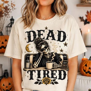 May include: A cream-colored t-shirt featuring a graphic of a skeleton with a messy bun and sunglasses sitting at a diner booth with a coffee mug. The text reads DEAD TIRED and Open 24/7. The design includes a crescent moon, stars, and a yellow rose.