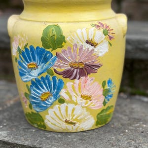 Puede incluir: Un jarrón de cerámica amarillo con dos asas laterales, adornado con flores pintadas a mano en tonos azules, blancos, rosas y morados. El diseño floral se asienta sobre un fondo amarillo, creando una pieza vibrante y decorativa.
