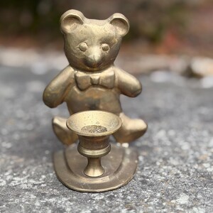 Può includere: Un portacandele a forma di orsetto in ottone con papillon. L'orso è seduto sopra una piccola coppetta per candele e una base rotonda. Il portacandele è in ottone e ha un aspetto vintage.