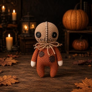 May include: A handmade crochet doll with a tan head and a stitched face featuring large black button eyes. The doll has an orange body with white cuffs and decorative patches. The background includes pumpkins, candles, and autumn leaves, creating a Halloween theme.
