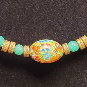 May include: A beaded necklace with a central oval bead featuring a colorful floral design. The necklace includes white round beads, gold-toned spacers, and turquoise-colored beads. The necklace is displayed on a black stand.