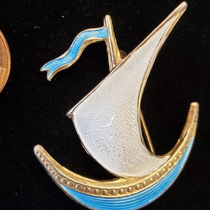 May include: A vintage sailboat brooch with a white sail, blue accents, and gold-tone metal. The brooch is next to a penny for size comparison. The brooch has a blue flag and a blue base.