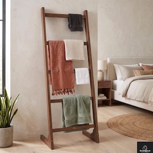 Peut inclure: Un porte-serviettes en bois dans une chambre. Le porte-serviettes est incliné contre un mur et contient des serviettes pliées de différentes couleurs, dont le gris, le rouille, le blanc et le vert. Un pot de plante et un lit sont visibles en arrière-plan. Hauteur : environ 180 cm.