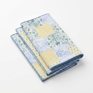 Puede incluir: Tres servilletas de tela plegadas con un diseño de patchwork. Las servilletas presentan estampados florales y de cuadros en azul, amarillo y blanco. Los bordes están ribeteados con un borde azul.