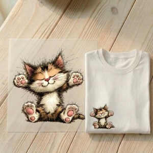 Könnte beinhalten: Ein weißes T-Shirt mit einer kleinen, farbenfrohen Grafik einer Cartoon-Katze mit ausgestreckten Armen. Neben dem Shirt befindet sich ein größerer Druck des gleichen Katzenmotivs. Die Katze ist braun, hellbraun und weiß.