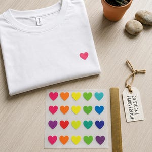 Könnte beinhalten: Ein weißes T-Shirt mit einem kleinen, auf die Brust gestickten rosa Herz, dazu ein Bogen mit zwanzig bunten Herzaufklebern, ein Metalllineal und ein Holzanhänger mit dem schwarzen Aufdruck 20 STÜCK | FARBVERLAUF.