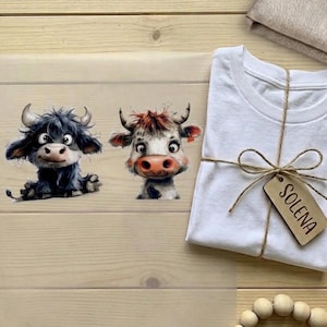 Könnte beinhalten: Ein weißes T-Shirt, das mit Bindfaden und einem Holzanhänger mit der Aufschrift "SOLENA" gebunden ist. Links sind zwei Cartoon-Kuh-Illustrationen zu sehen. Eine ist schwarz und die andere weiß und braun. Der Hintergrund ist helles Holz.