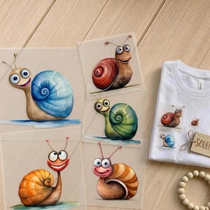 Könnte beinhalten: Ein weißes T-Shirt und mehrere illustrierte Karten mit Cartoon-Schnecken mit bunten Gehäusen. Die Schnecken haben ausdrucksstarke Gesichter und sind in verschiedenen Farben dargestellt, darunter Blau, Rot, Orange und Grün. Ein Holzschild mit dem Namen "SOLENA" ist ebenfalls sichtbar.