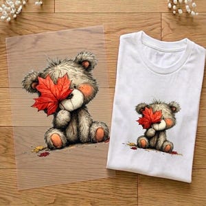 Könnte beinhalten: Ein weißes T-Shirt und eine transparente Folie, beide mit einer grafischen Illustration eines flauschigen grauen Teddybären, der sitzt und ein leuchtend rotes Ahornblatt vor sein Gesicht hält. Der Bär hat orangefarbene Wangen und Tatzen.