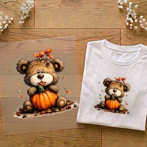 Könnte beinhalten: Ein weißes T-Shirt mit einer grafischen Illustration eines braunen Teddybären, der einen orangefarbenen Kürbis hält, umgeben von verstreuten Herbstblättern. Das gleiche Bären- und Kürbismotiv ist auf einer transparenten Folie daneben abgebildet.