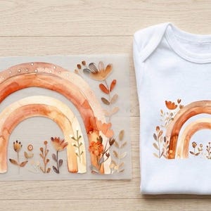 Könnte beinhalten: Ein weißer Baby-Body mit einem Aquarell-Regenbogen-Design in Orange- und Brauntönen, umgeben von floralen Elementen. Daneben befindet sich eine Transferfolie mit dem gleichen Regenbogen- und Blumendesign.