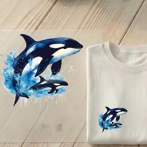 Könnte beinhalten: Ein weißes T-Shirt mit einem Aquarelldesign von zwei Orcas, die aus dem Wasser springen. Die Orcas sind schwarz und weiß mit blauen Wasserspritzern. Ein ähnliches Design ist auf einer transparenten Folie gedruckt.