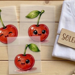 Könnte beinhalten: Ein weißes T-Shirt mit einem Holzanhänger mit der Aufschrift "SOLENA", der mit Bindfaden gebunden ist. Das Bild zeigt auch drei Illustrationen von lächelnden Kirschen mit grünen Blättern auf hellem Hintergrund.