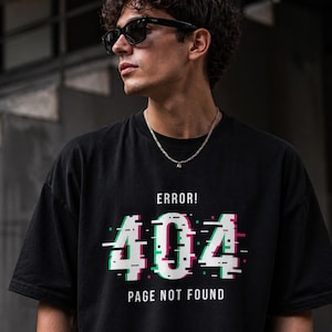 May include: Black t-shirt with the text "ERROR! 404 PAGE NOT FOUND" in a glitch effect design. The graphic features white, green, and pink colours. The shirt is worn by a person wearing sunglasses and a silver chain.