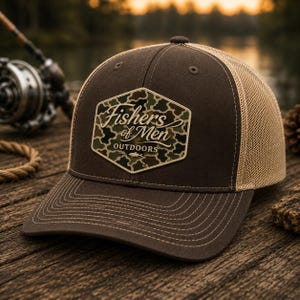 May include: A brown baseball cap with a tan mesh back and a hexagonal patch on the front. The patch features a camouflage pattern with the text Fishers of Men OUTDOORS and a small fish icon. The cap has a curved brim with visible stitching.