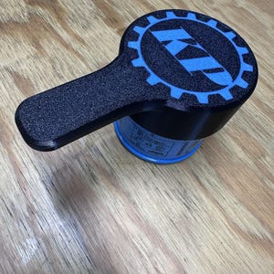 May include: A black oil filter wrench with a textured finish and a long handle. The top of the tool features a blue gear-shaped design with the letters KP printed in the center. The wrench is attached to a blue cylindrical oil filter.