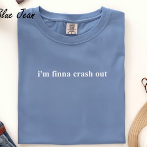 May include: A light blue t-shirt with the text "i'm finna crash out" in white. The shirt is folded and has a Comfort Colors tag. The shirt is on a white surface with a pair of glasses and a hat.