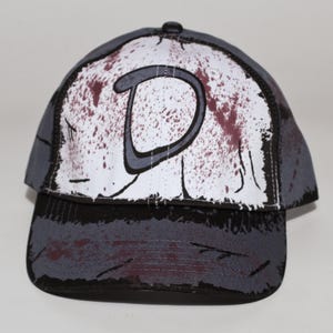 May include: A baseball cap with a gray and black brim and a white front panel. The front panel features a large gray letter "D" with a maroon splatter design. The cap has a black button on top.